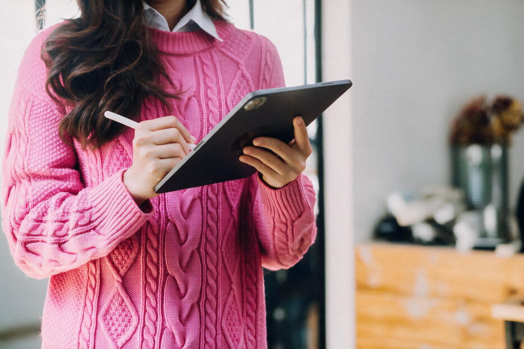 Woman in a bright pink sweater standing in her home using a stylus on a tablet.